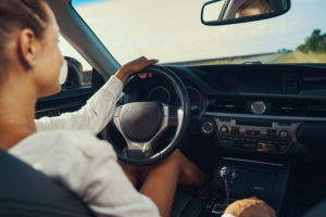 Vrouwelijke bestuurder in een auto, gezien van achteren, terwijl ze met één hand het stuur vasthoudt en ontspannen over een lege weg rijdt op een zonnige dag.