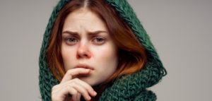 Jonge vrouw met een bezorgde blik, rode ogen en neus, gekleed in een dikke groene sjaal met capuchon.